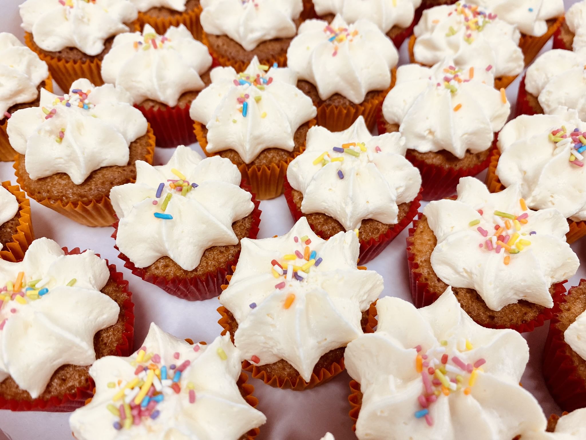 A close-up view of multiple mini vanilla cupcakes topped with rich vanilla buttercream frosting and colorful sprinkles. The cupcakes are arranged closely together, creating a festive and indulgent display.